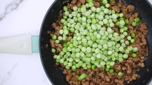 Close-up of frozen peas being poured into a pan of cooked ground beef.