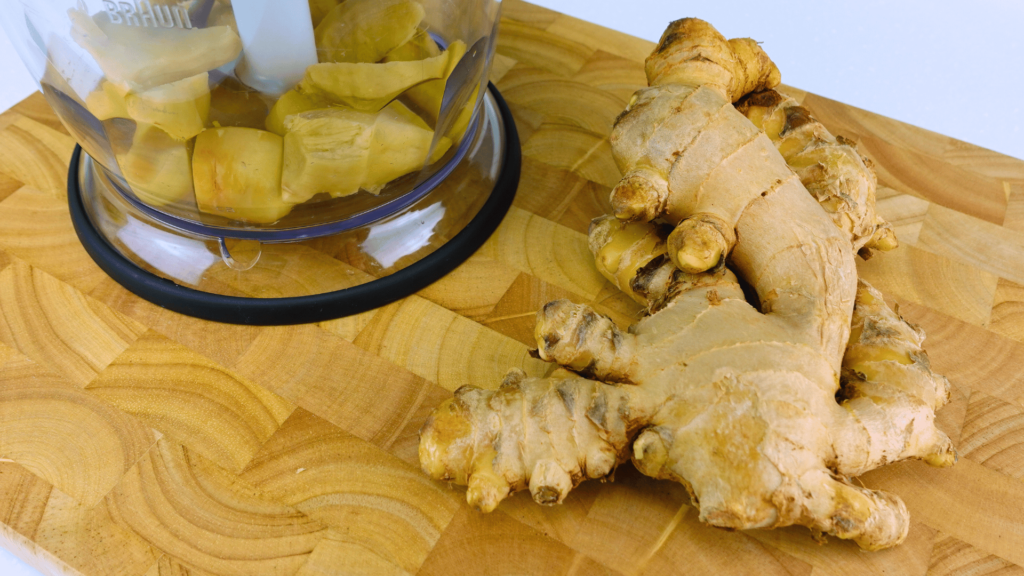 Peeled ginger cut into cubes in a blender bowl with unpeeled ginger nearby.